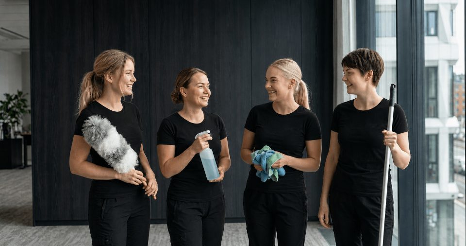 Four smiling cleaners in black t-shirts holding a duster, a spray bottle, cloths and a mop
